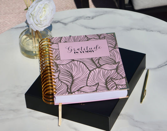 Soft pastel flat-lay of the physical Gratitude is Yummy Journal with gold spiral binding, pink cover, gold pen, and white ros