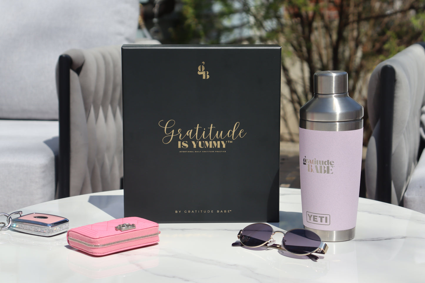 Gratitude is Yummy Journal product display with 'Gratitude is Yummy' matte black gift box with gold foil writing, pink wallet, sunglasses, and YETI branded water bottle on a table outdoors.