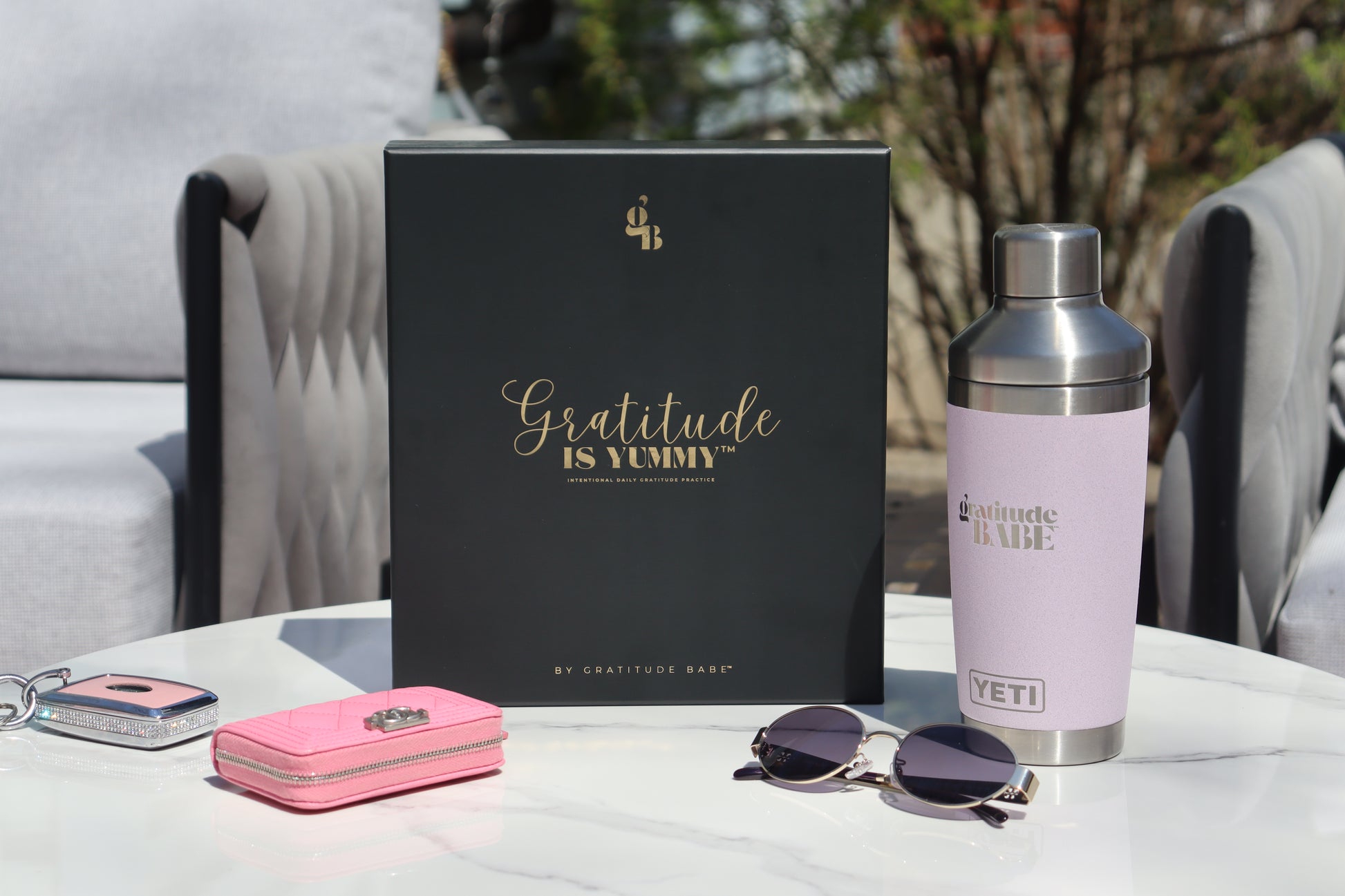 Gratitude is Yummy Journal product display with 'Gratitude is Yummy' matte black gift box with gold foil writing, pink wallet, sunglasses, and YETI branded water bottle on a table outdoors.