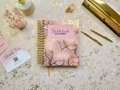Pink and gold notebook with 'Gratitude is yummy' on a textured surface with stationery items.