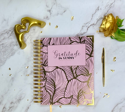 Pink and gold notebook with 'Gratitude is Yummy' on a marble surface with decorative elements.