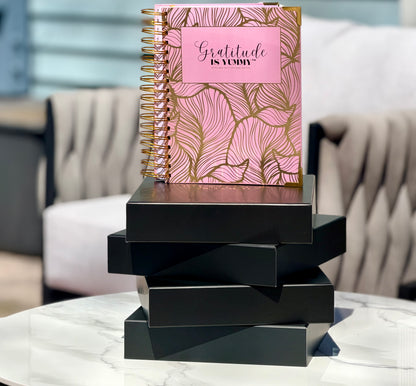Blush pink spiral journal with gold foil, with 'Gratitude is Yummy' on a a stack of black gift boxes. Pink. Girly. Aesthetic. Mindset. Manifestation. outdoor. Gratitude journal. Manifestation journal. Gratitude manifestation journal. 