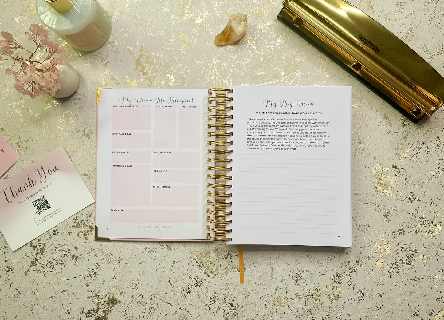 Open planner with pink and gold design on a decorative surface 

Open spiral-bound planner on a textured surface with stationery items around