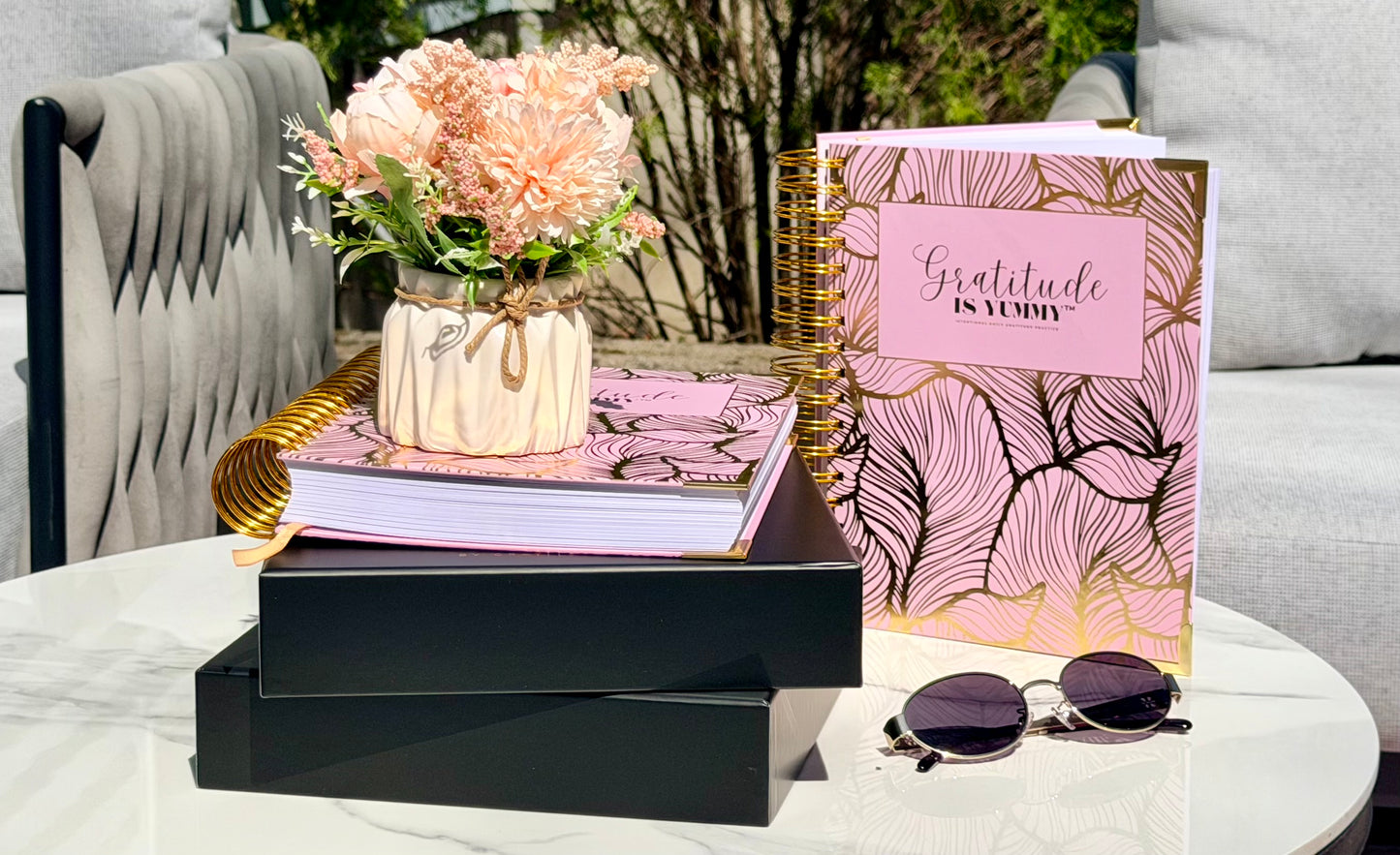 Decorative table setting with Blush pink spiral journals with gold foil, with 'Gratitude is Yummy' on a a stack of black gift boxes. Pink. Girly. Aesthetic. Mindset. Manifestation. outdoor. Gratitude journal. Manifestation journal. Gratitude manifestation journal. Spiral books, a vase of flowers, and Tory Burch sunglasses on a white surface.