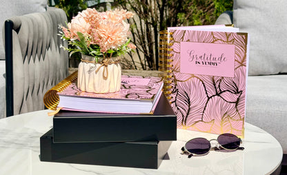 Decorative table setting with Blush pink spiral journals with gold foil, with 'Gratitude is Yummy' on a a stack of black gift boxes. Pink. Girly. Aesthetic. Mindset. Manifestation. outdoor. Gratitude journal. Manifestation journal. Gratitude manifestation journal. Spiral books, a vase of flowers, and Tory Burch sunglasses on a white surface.