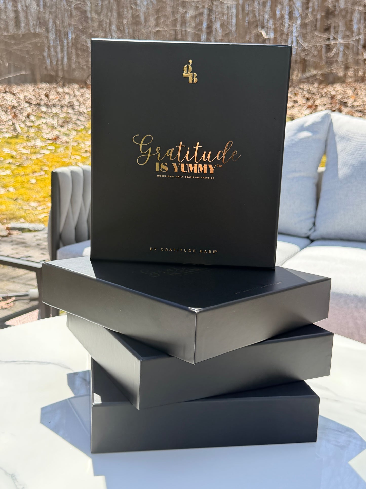 Stack of black gift boxes with gold foil, with 'Gratitude is Yummy' on a marble surface outdoors. Girly. Aesthetic. Mindset. Manifestation. Gratitude journal. Manifestation journal. Gratitude manifestation journal. Black gift boxes with gold text on a textured surface.