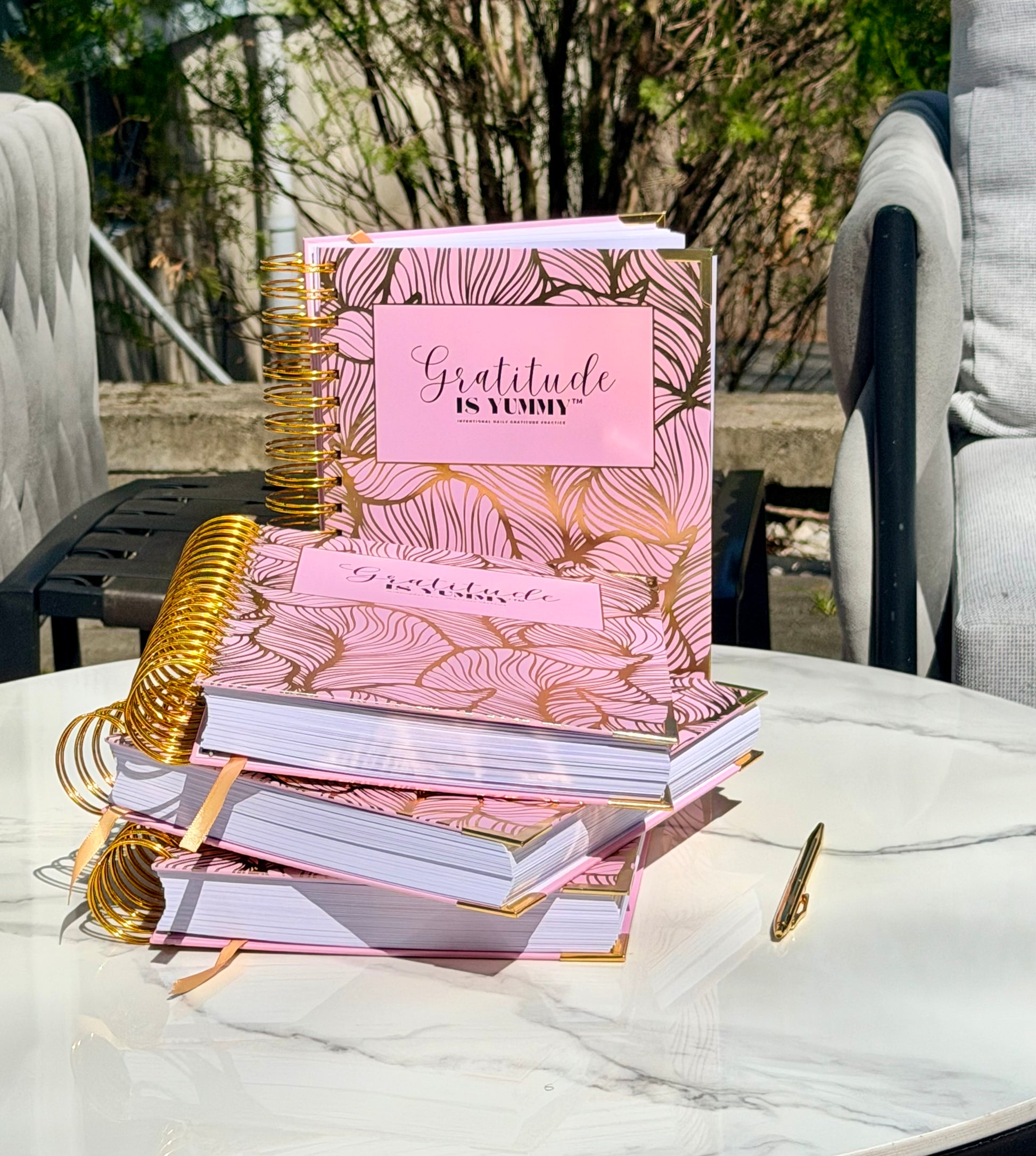 Stack of Blush pink spiral journal with gold foil, with 'Gratitude is Yummy' on a marble surface outdoor with a gratitude babe engraved gold pen. Pink. Girly. Aesthetic. Mindset. Manifestation. Outdoor. Gratitude journal. Manifestation journal. Gratitude manifestation journal