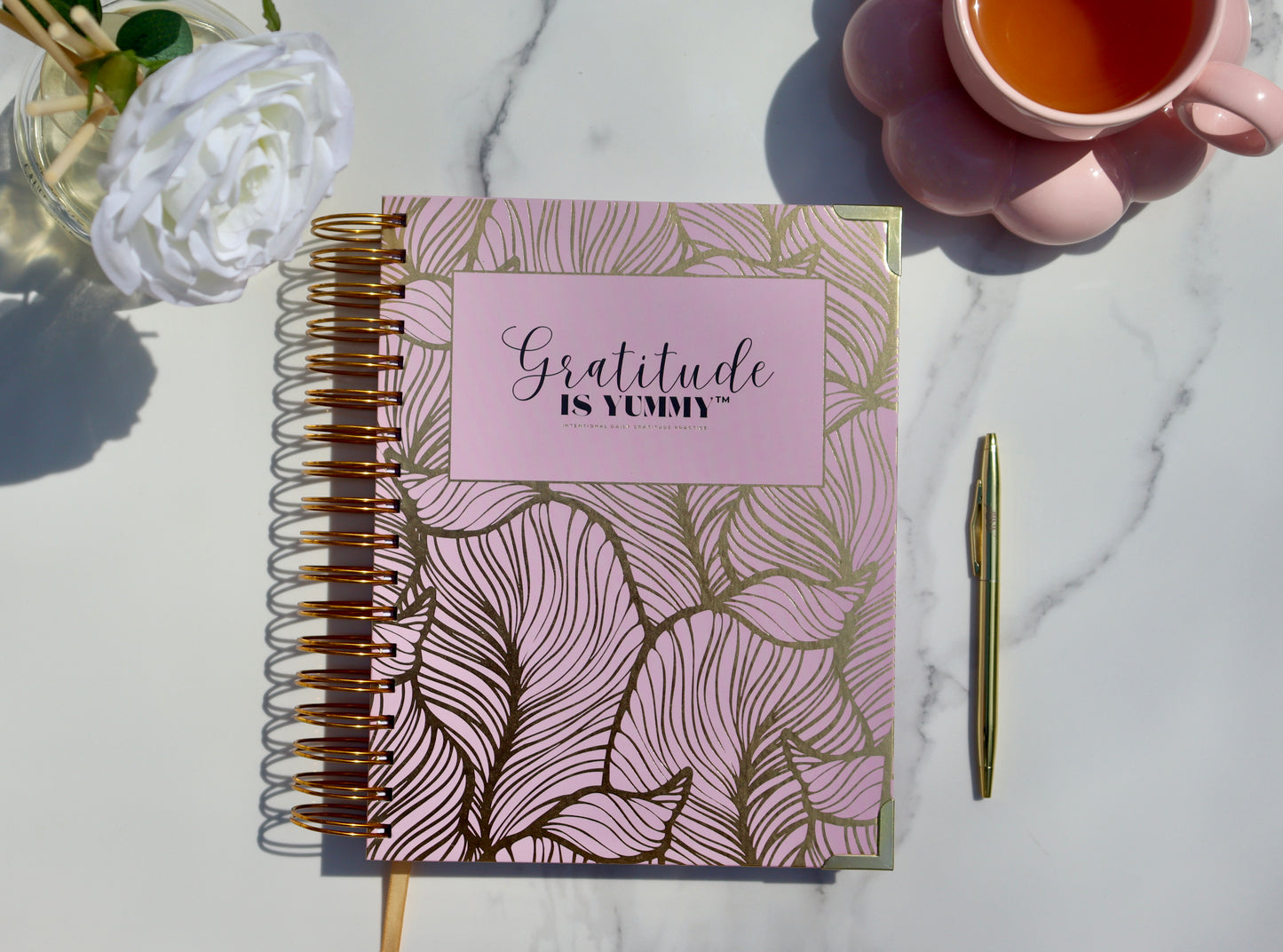 Blush pink spiral journal with gold foil, with 'Gratitude is Yummy' on a marble surface with a cup of tea and gratitude babe engraved gold pen. Pink. Girly. Aesthetic. Mindset. Manifestation