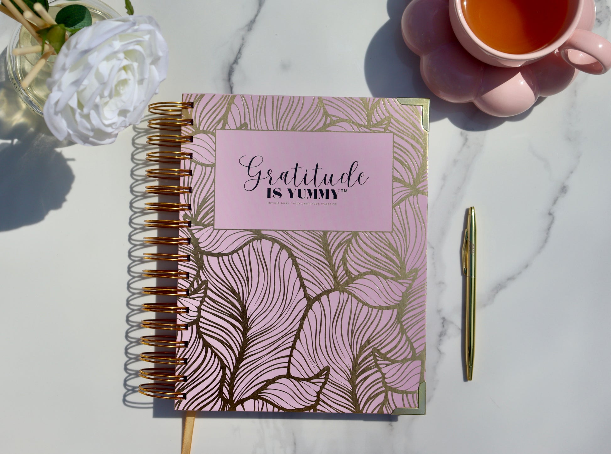 Blush pink spiral journal with gold foil, with 'Gratitude is Yummy' on a marble surface with a cup of tea and gratitude babe engraved gold pen. Pink. Girly. Aesthetic. Mindset. Manifestation
