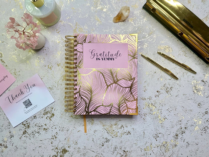 Pink and gold notebook with 'Gratitude is yummy' on a textured surface with stationery items.
